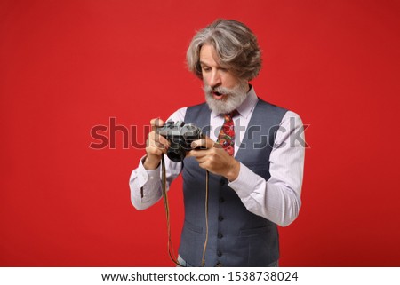Amazed elderly gray-haired mustache bearded man in classic shirt vest colorful tie isolated on red background in studio. People lifestyle concept. Mock up copy space. Hold retro vintage photo camera