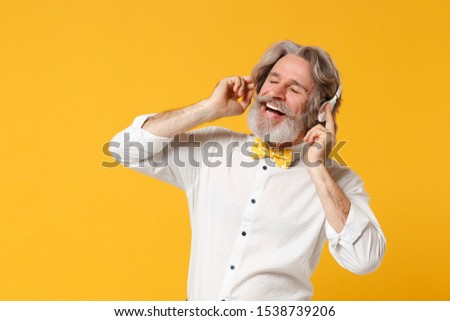 Relaxed elderly gray-haired mustache bearded man in white shirt bow tie isolated on yellow background, studio portrait. People lifestyle concept. Mock up copy space. Listening music with headphones