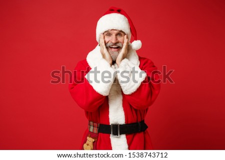 Funny elderly gray-haired mustache bearded Santa man in Christmas hat posing isolated on red wall background. Happy New Year 2020 celebration holiday concept. Mock up copy space. Put hands on cheeks