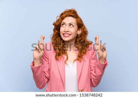 Redhead woman in suit over isolated blue wall with fingers crossing and wishing the best