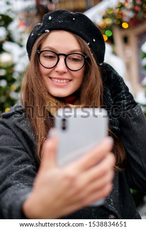 Beautiful young woman taking a selfie with christmas tree behind her with mobile phone camera
