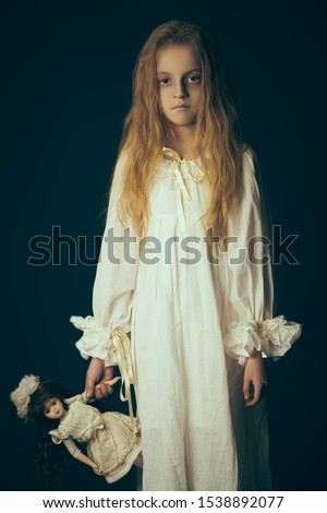Scary little girl ghost in a white nightgown holds her doll. Black background. Halloween.  Royalty-Free Stock Photo #1538892077