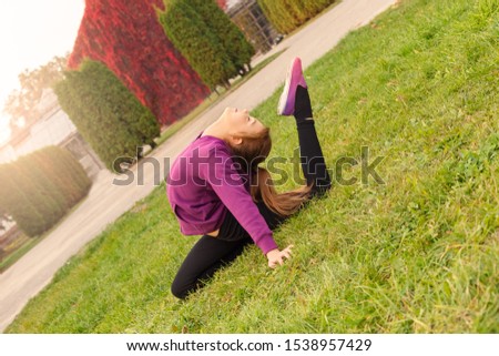 Little girl gymnast sitting on grass doing exercise bending back to bent leg flexible outdoors in the autumn park closed eyes smiling joyful