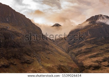 One of the three mountains called The Three Sisters, covered in clouds and brown wintery vegetation on a partially cloudy day in Scottish Highlands.