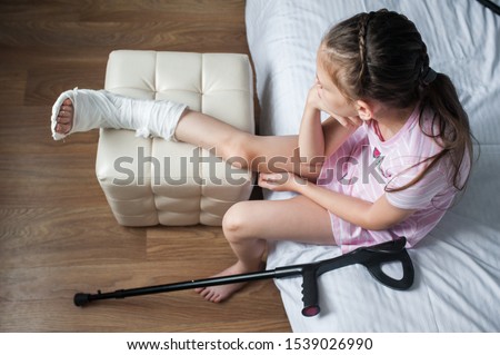 Girl child with a broken leg in a cast is sitting on a bed with crutches.
