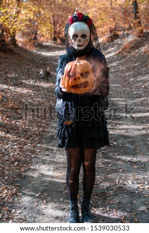 Photo of  walking girl with creepy skull mask in the autumn woods, holding a carved smoky Halloween pumpkin.  On background path in autumn forest with fall leaves. Concept about Halloween.
