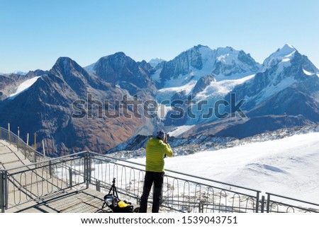 A tourist taking photos on the platform of Corvatsch Cableway mountain station with a stunning view of Piz Bernina & glaciers among Swiss Alps under blue clear sky near St Moritz, Grisons, Switzerland