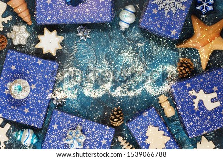 2020 in glitter, Chritmas background. Presents in blue wrapping paper with silver sparkles, wooden decorations, ornaments on blue table, copy space in the middle, top view, selective focus