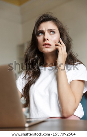 Photo of a confused young woman indoors at home using laptop computer talking by mobile phone.