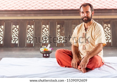 Thai massage teacher and therapist ready to teach students traditional nuad boran techniques. Man sit on white yoga mat on red trouses. Flower and instruments on background. Yoga coaching instructor. Royalty-Free Stock Photo #1539092609