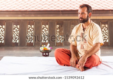 Thai massage teacher and therapist ready to teach students traditional nuad boran techniques. Man sit on white yoga mat on red trouses. Flower and instruments on background. Yoga coaching instructor. Royalty-Free Stock Photo #1539092612