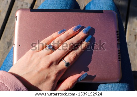 Young woman holding a pink laptop in her lap. Baby blue sparkly manicured nails and rings. Accesories close up and using technology on the go. Getting work done in the outdoor office.