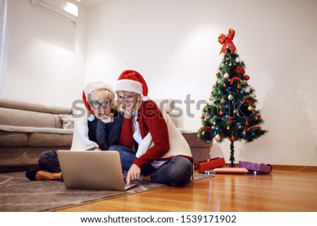 Beautiful Caucasian blonde woman sitting with her mother on floor in living room and using laptop. Both having santa hats on heads. In background is decorated christmas tree.