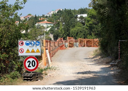 Road under construction with speed limit and warning road signs at start of construction site surrounded with protective fence and dense trees with family houses and clear blue sky in background