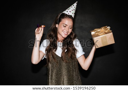 Image of nice smiling woman in party cone holding gift box and pipe while dancing isolated over black wall