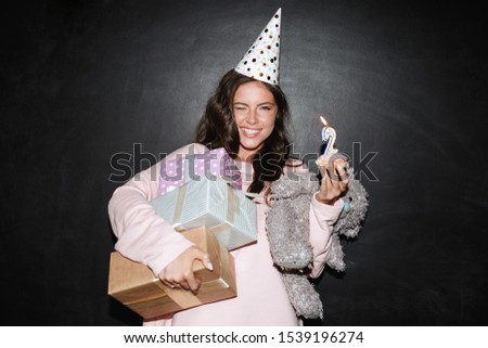 Image of smiling woman in party cone holding gift boxes and donut with candle while winking isolated over black wall