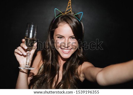 Image of cute joyful woman in toy ears holding glass of champagne while taking selfie photo isolated over black wall