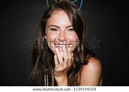 Image of brunette smiling woman in toy ears holding glass of champagne while laughing isolated over black wall