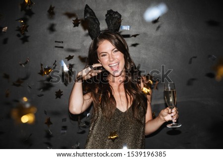 Image of brunette happy woman in bunny ears holding glass of champagne while dancing isolated over black wall