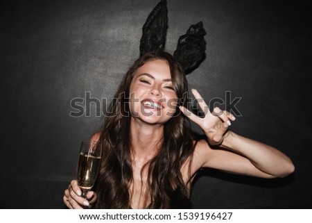 Image of brunette smiling woman in bunny ears holding glass of champagne while gesturing peace sign isolated over black wall