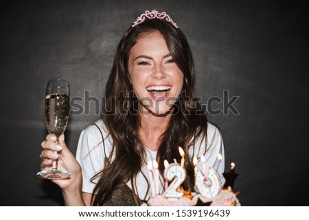Image of brunette excited woman in tiara holding cakes and glass of champagne while laughing isolated over black wall