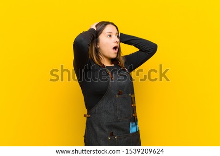 hairdresser woman with open mouth, looking horrified and shocked because of a terrible mistake, raising hands to head against orange background