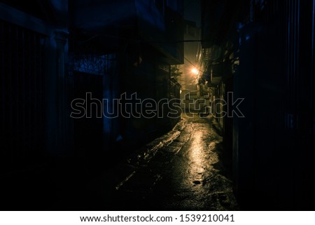 A dark, shadowy and dangerous looking urban back-alley at night time in suburbs Hanoi, Vietnam. Low light reflected on wet pavement from post lamp at the end of long road corner Royalty-Free Stock Photo #1539210041