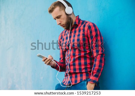 A handsome man holds a phone in his hands and listens to music on headphones. He wears a colorful shirt and stands in front of a blue wall