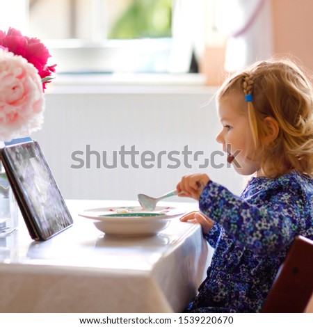 Adorable toddler girl eating healthy cereal with milk for breakfast and watching cartoons on tablet pc. Cute happy baby child in colorful clothes sitting in kitchen