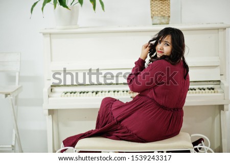 Attractive south asian woman in deep red gown dress posed at room and play on piano.