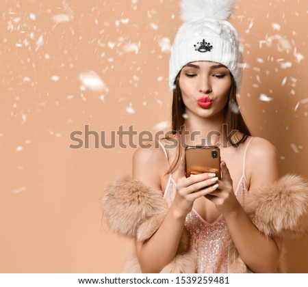 Happy New Year and Merry Christmas. Young woman in shimmering cocktail gown, fur coat and warm hat is sending a kiss to her friends family lover by cell phone under the snow