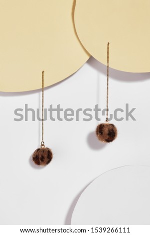 Subject shot of stud earrings in the form of the brown fur balls with leopard spots dangling on the long golden chains. The earrings are fixed on the beige circular plates on the white background. 