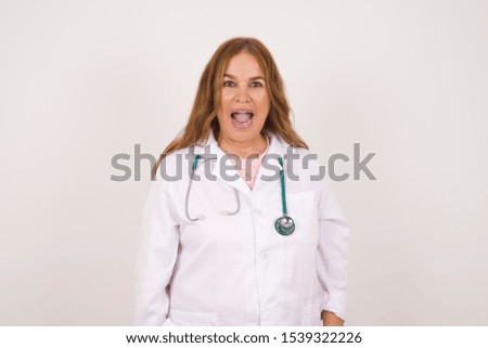 Emotive doctor woman wears medical uniform, happy and excited to celebrate anniversary. Happy young woman has good mood stands against gray wall.