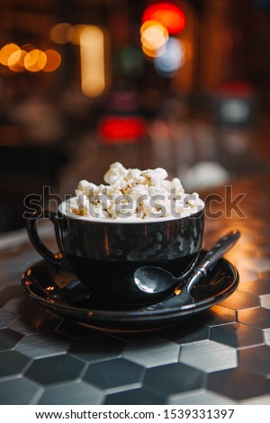 Mug of cappuccino coffee with popcorn on a bar counter in the light of colored lamps. The cup is black. Mosaic table. Cocoa. Great background for Photoshop, suitable in the menu.