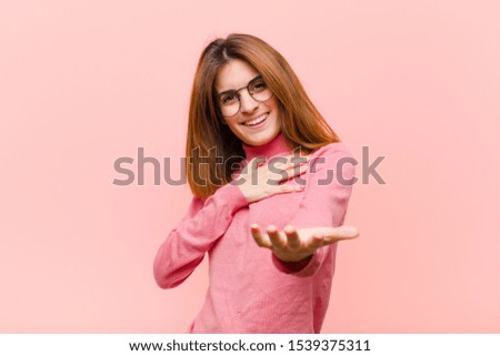 young pretty woman feeling happy and in love, smiling with one hand next to heart and the other stretched up front against pink background