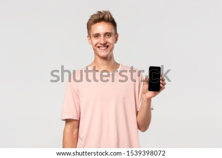Smiling blond teenager in a pink tee pointing at the screen of a smartphone isolated over light grey background. Place for advertising