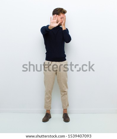 young full body man covering face with hand and putting other hand up front to stop camera, refusing photos or pictures against white background