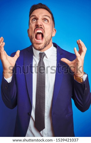 Vertical angle picture of young handsome business man over blue isolated background crazy and mad shouting and yelling with aggressive expression and arms raised. Frustration concept.