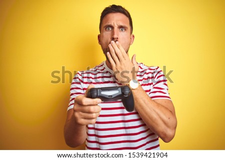 Young handsome man playing video games using joystick game pad over yellow background cover mouth with hand shocked with shame for mistake, expression of fear, scared in silence, secret concept