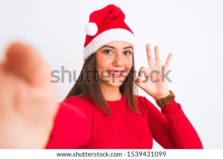 Girl wearing Christmas Santa hat make selfie by camera over isolated white background doing ok sign with fingers, excellent symbol