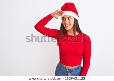 Young beautiful girl wearing Christmas Santa hat standing over isolated white background very happy and smiling looking far away with hand over head. Searching concept.