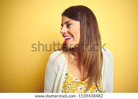 Young beautiful woman wearing jacket standing over yellow isolated background looking away to side with smile on face, natural expression. Laughing confident.