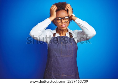 Young african american woman shop owner wearing business apron over blue background suffering from headache desperate and stressed because pain and migraine. Hands on head.