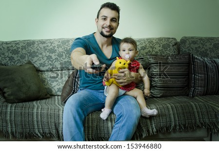 Father holds baby with pacifier and watching tv with remote control.