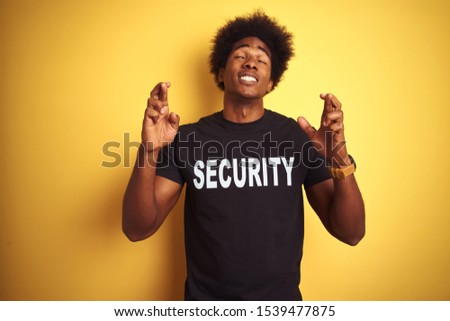 American safeguard man with afro hair wearing security uniform over isolated yellow background gesturing finger crossed smiling with hope and eyes closed. Luck and superstitious concept.