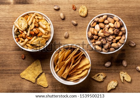 Various beer snacks in white bowls on a wooden table, top view, flat lay. Nachos, pistachios, cheese sticks and other beer snacks on a wooden background