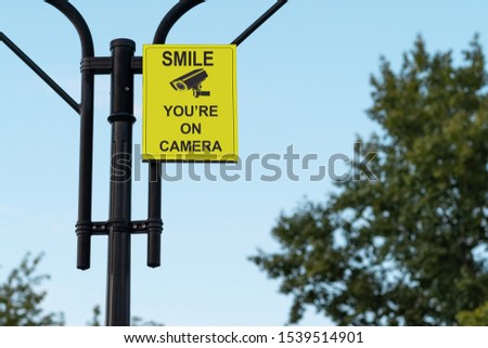 
Smile, you're on camera. Yellow sign on a metal pillar against a blue sky and treetops in a park. Warning sign