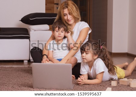 Young mother and her children are using a laptop and smiling while lying together on the floor at home. Family with laptop surfing in the net while choosing movie or cartoons to watch at leisure.