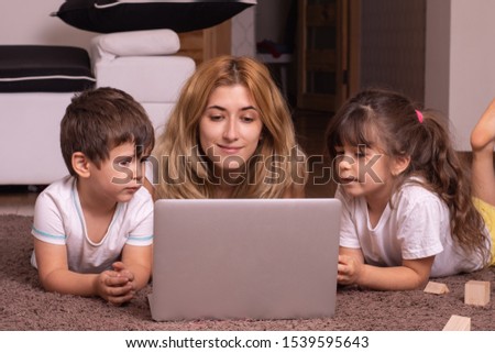 Young mother and her children are using a laptop and smiling while lying together on the floor at home. Family with laptop surfing in the net while choosing movie or cartoons to watch at leisure.
