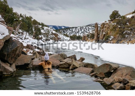 CLOSE UP: Young woman hiking in Colorado relaxes in the Radium Hot springs. Girl in a black bikini and white winter cap lies in the therapeutic water of ahot pond and observes the snowy nature. Royalty-Free Stock Photo #1539628706